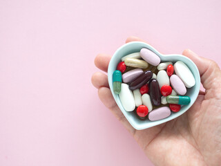 Hand holding a heart-shaped blue bowl filled with various colorful pills and capsules over pink...