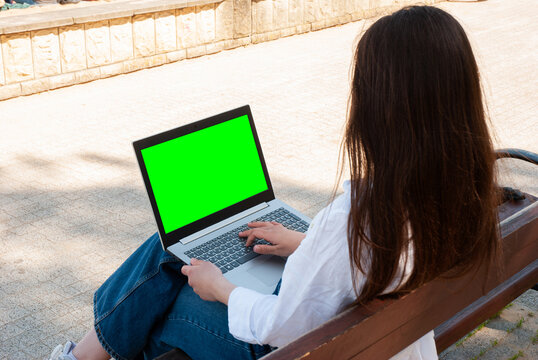 Young caucasian female using laptop with green screen outdoors on a sunny day, sitting on a wooden bench in casual attire, rear view. Digital nomad lifestyle. Remote work