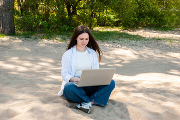 Caucasian young adult female in casual attire focuses on laptop while seated on a sunny sandy beach...