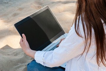 Young female working remotely on a laptop while sitting on sandy beach, embracing the summer sun and blending work with relaxation in a casual setting. Digital nomad lifestyle. Remote work