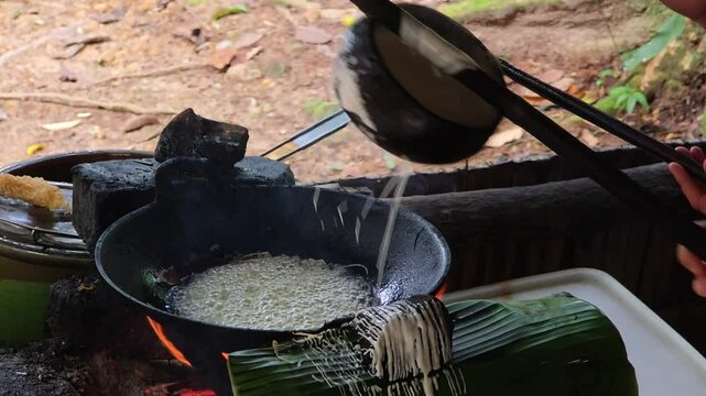 Hands expertly pour batter to create threads of traditional Kuih Jala (Jala Emas) snack, frying over fire on a banana leaf at Mari Mari village in Malaysia
