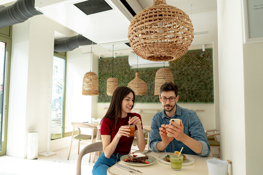 Happy couple enjoying a meal while using a smartphone in a trendy cafe, savoring brunch and sharing smiles in a vibrant atmosphere - Powered by Adobe