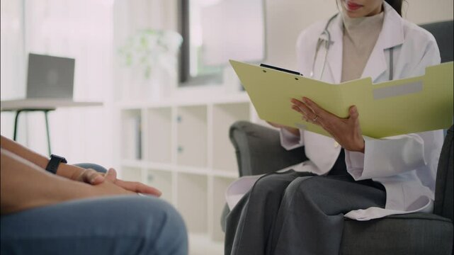 Asian female doctor explaining health report to elderly patient at clinic. Medical consultation with document and yellow folder in professional.