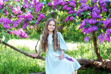 Happy portrait of a beautiful girl with long hair and in dress sitting near a blossoming lilac....