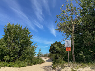 A sandy path leading towards the beach, marked by a sign saying „ENTRANCE 6 PIASKI," under a vibrant blue sky, Baltic Sea coast, Poland.