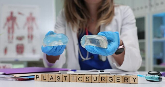 Female surgeon wearing blue gloves displays silicone breast implants against phrase Plastic Surgery on wooden blocks. Concept of cosmetic enhancement