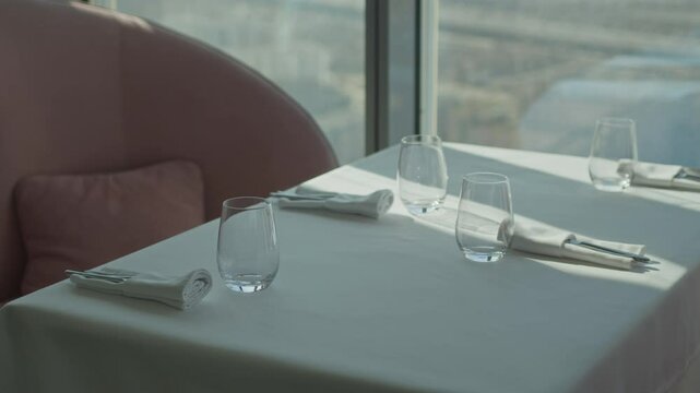 No people shot of dining table set with white tablecloth, four transparent glasses for beverages and cutlery in folded napkins in modern restaurant with panoramic view from windows
