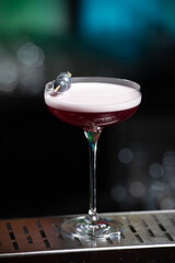 purple cocktail with a foamy white top is garnished with skewered blueberries on a textured bar surface