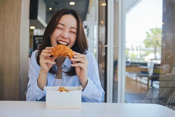 A joyful young Asian woman enjoying a piece of fried chicken at a modern restaurant. She laughs wholeheartedly, holding the crispy food.