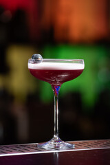 purple cocktail with a foamy white top is garnished with skewered blueberries on a textured bar surface