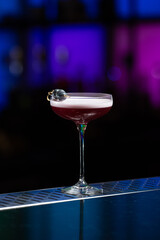 purple cocktail with a foamy white top is garnished with skewered blueberries on a textured bar surface