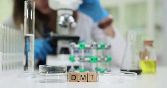 Letters DMT on wooden blocks displaying drug and technician with microscope in laboratory. Female researcher conducts scientific investigation in lab