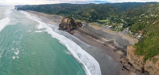 Aerial view of Piha Beach, New Zealand, featuring Lion Rock. Waves crash on the black sand beach, popular for surfing and scenic views. Lush green hills surround the coastline. PIHA, NZ