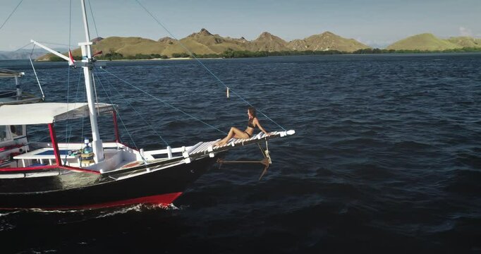 Aerial drone footage of a young tourist relaxing and sunbathing on the bowsprit of a yacht sailing in the beautiful Komodo National Park, Indonesia