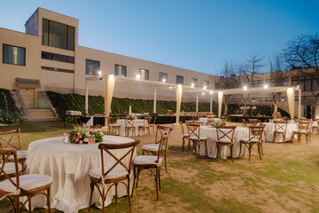 Outdoor event space featuring round tables with floral centerpieces and wooden chairs under a canopy, set against a modern building and twilight sky.