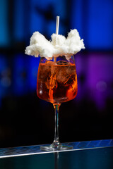 A vibrant orange cocktail filled with ice sits on a textured bar, topped with a whimsical cloud of white cotton candy and a white straw.