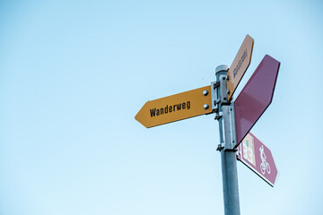 Signpost displaying directional arrows, including a yellow Wanderweg sign, set against a bright blue sky, representing outdoor adventure and navigation choices