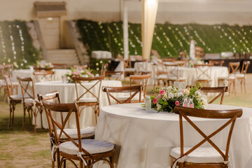 Elegantly styled outdoor wedding reception featuring round tables, wooden cross-back chairs, and lush floral centerpieces under soft, romantic lighting.