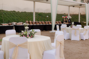 Elegant outdoor event with round tables adorned with flowers and white chairs with bows. Buffet line is visible.