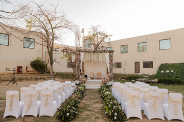 Outdoor wedding setup with white chairs, floral aisle, and a decorated altar against a building backdrop, ready for a celebration of love and union.