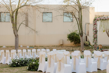 Outdoor wedding setup with white chairs adorned with ribbons, floral decorations, and a decorated archway in a serene garden setting.
