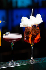 Two vibrant cocktails sit on a bar counter, one a refreshing green mix with mint and a straw, the other a bright orange topped with white sugar cotton candy and a straw.