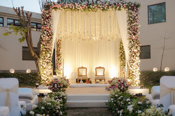 Elaborate outdoor ceremony stage. Floral arrangements with white chairs & curtains. Wedding decor with a rustic & elegant vibe. Soft lighting for ambiance.