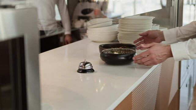 Cropped shot of unrecognizable restaurant kitchen worker ringing bell on counter passing ordered fine dining dishes to server in classic uniform