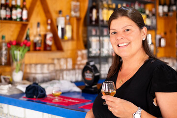 Smiling Woman Holding Whisky Glass at Bar – Happy Female Enjoying Drink in Cozy Pub Setting with Bottles and Flowers in Background, Relaxed Social Atmosphere
