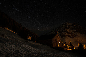 Panoramic night view of the starry sky, Milky Way, and an illuminated stone building on the snow in the foreground.