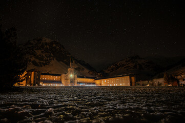 Panoramic night view of the starry sky, Milky Way, and an illuminated stone building on the snow in the foreground.