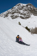 Father and daughter playing and enjoying a snowy day in the mountains, sliding it down the slope.