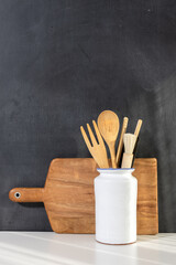 Wooden kitchen utensils in white ceramic container and wooden cutting board leaning against a blackboard wall with copy space, creating a cozy kitchen scene