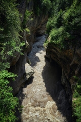 Wide river rushes through a steep, rocky gorge, flanked by vibrant green foliage, under bright sunlight. The natural beauty captivates with its untamed landscape