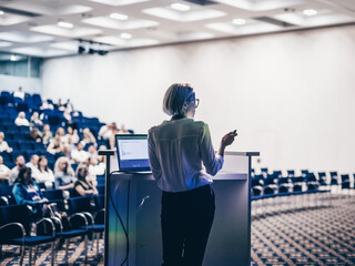 Female speaker giving a talk on corporate business conference. Unrecognizable people in audience at...