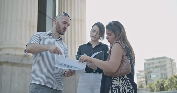 Three business colleagues review documents outside a courthouse on a summer day. They discuss and make notes on the paperwork.