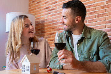 Smiling couple toasting with red wine while celebrating their new home. A miniature house and keys on the table symbolize the beginning of a new chapter in their life together. 