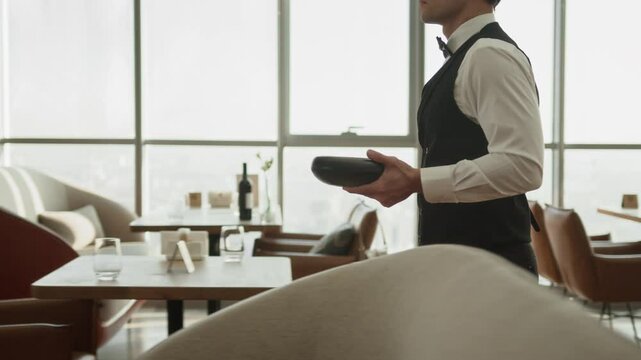 Cropped tracking shot of server in elegant classic unifrom bringing ordered meals in bowls working in fine dining restaurant with panoramic windows