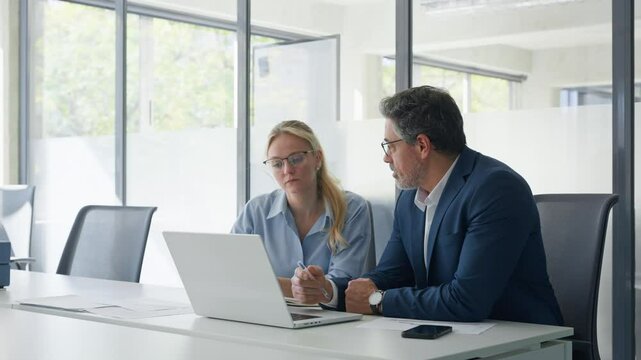 Mature focused senior mentor leader man showing young woman manager project on laptop in modern office. Two partners, team of professional business people working, studying, writing notes sit at desk