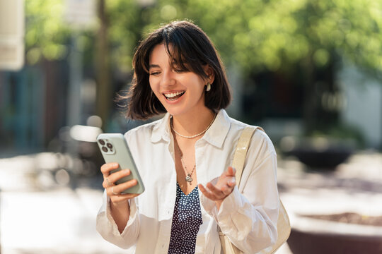 Sharing the happiness and good news. Young attractive woman smiling while using phone video chat or browsing, smiling in the city on a sunny summer day