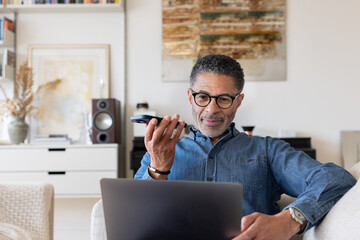 Mature man Using Voice Assistant whilst working on a laptop at home
