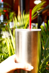 Hand holding travel mug with blurred green leaves background in sunny day. Close-up tumblers for cold drink in summer. Concept of lifestyle, eco-friendly, nature, destination, natural light, and leaf.