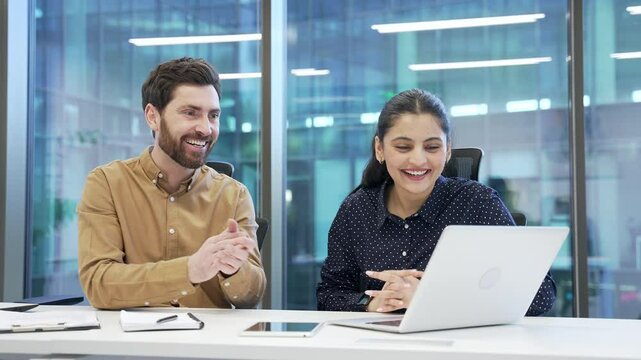 Two satisfied happy co-workers celebrate success at work using laptop. Smiling glad colleagues, a man and a woman, read good news on the computer sitting at desk at workplace in modern business office