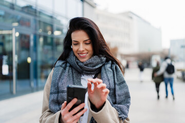 Smiling businesswoman in a stylish coat and scarf taking a selfie with her smartphone in front of a...