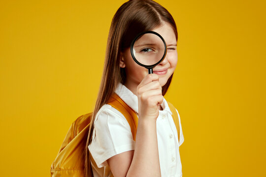 Inquisitive child with a backpack peeks through a magnifying glass, displaying a playful and curious demeanor against a yellow classroom background. Ideal for educational content and school promotions - Powered by Adobe