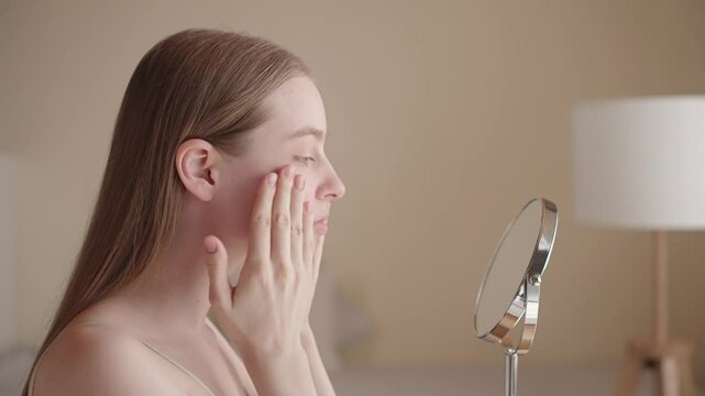 Young woman applying serum to her face during morning skincare routine. Natural beauty, clean skin, healthy lifestyle concept. Gen Z female gently massaging serum into her cheeks in front of mirror.