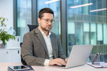 A serious young businessman in a suit is sitting at a desk in the office and working intently on a...
