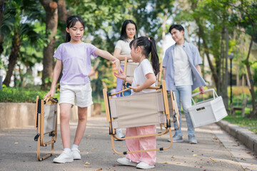 Children and adults enjoy a sunny day in the park carrying folding chairs while sharing smiles and...