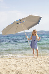 Cute little girl in blue summer dress under blue parasol on the empty senf beach with clear blue...