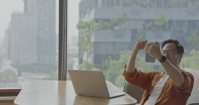 Senior Asian businessman wearing headset celebrates success during online meeting in modern co-working space. Remote work and business communication concept.